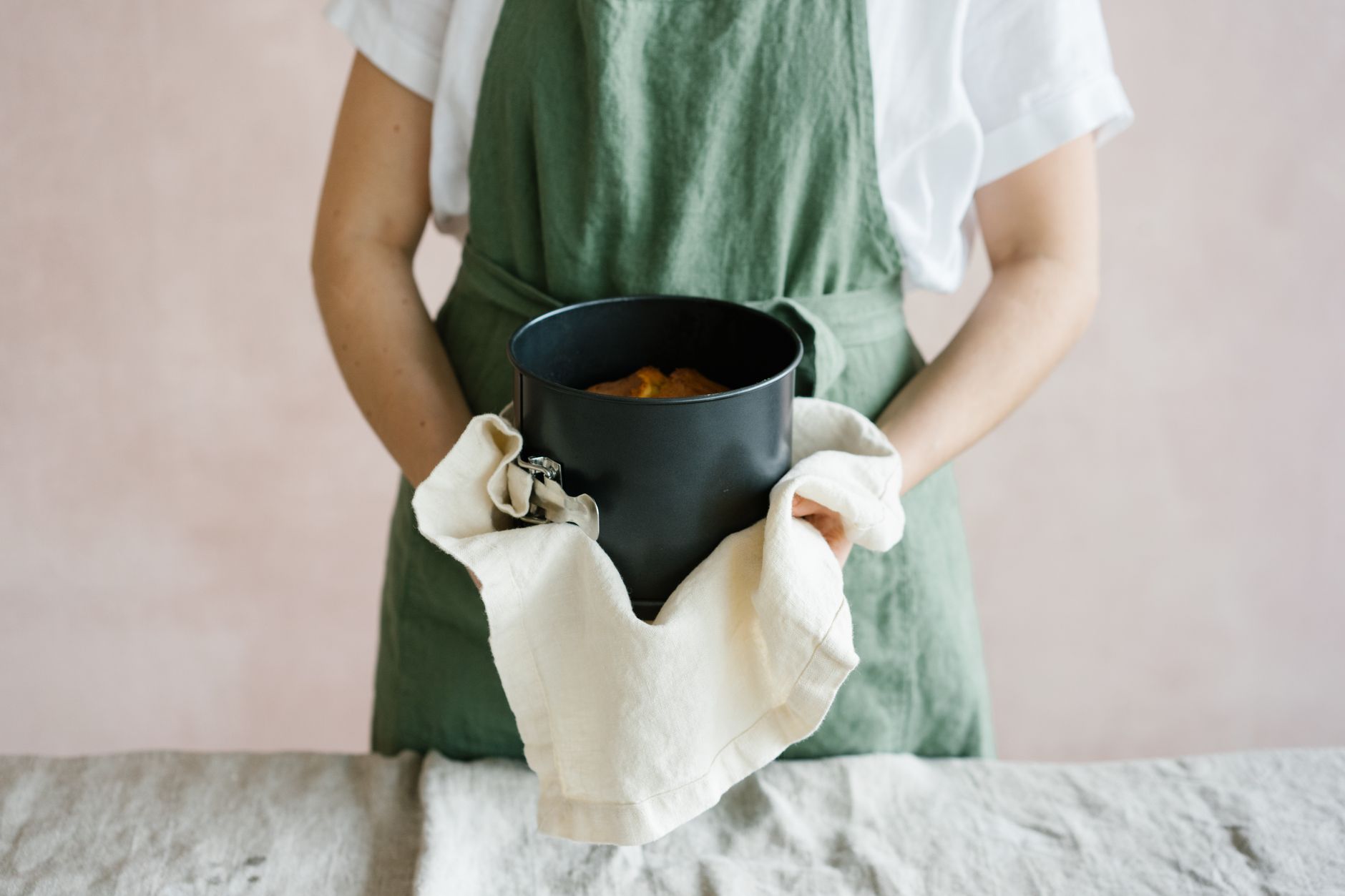 A person in an apron holding a round cake pan with a baked cake inside